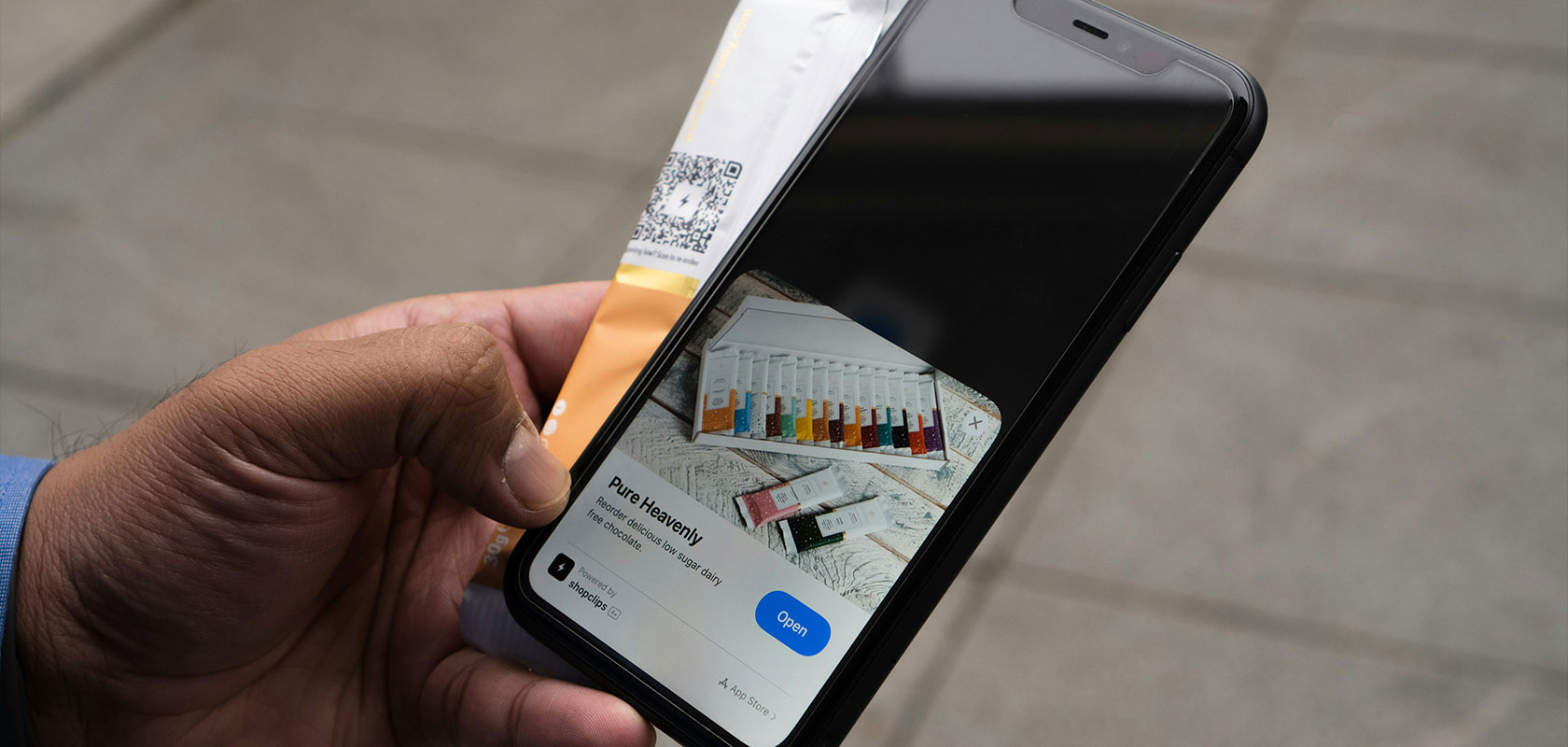 Eine Hand hält ein Smartphone, das eine App-Clip-Produktkarte der Marke „Pure Heavenly“ für Schokolade anzeigt, neben einer Verpackung mit QR-Code.