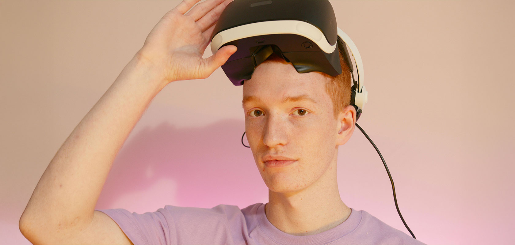 Person mit Virtual-Reality-Headset, die das Visier anhebt, mit neutralem Gesichtsausdruck. Der Hintergrund ist sanft in Rosa beleuchtet.