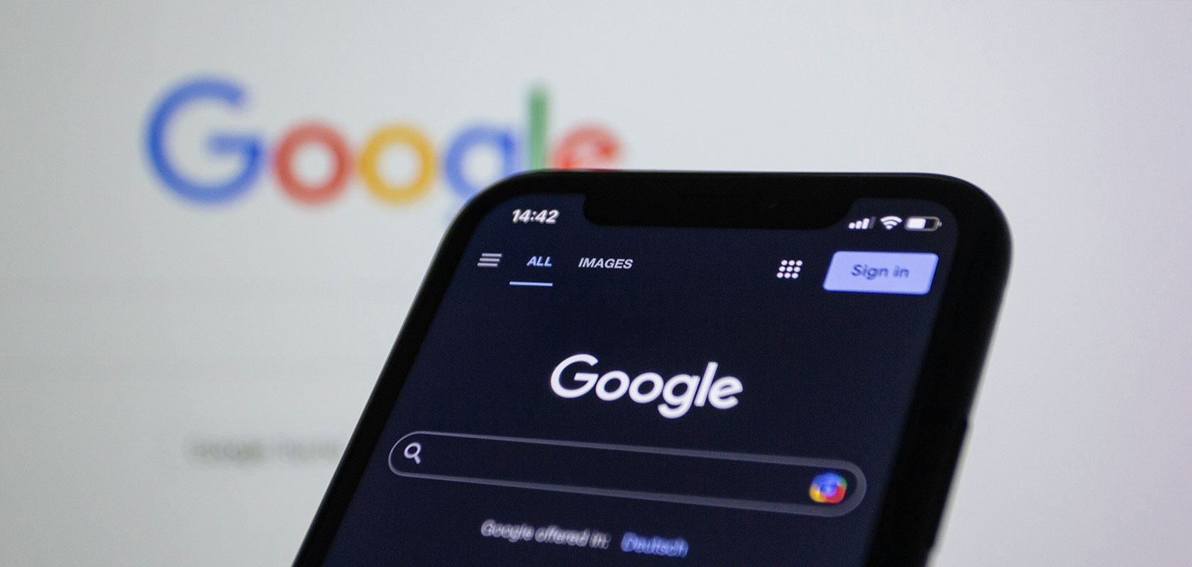 Smartphone im Vordergrund mit geöffneter Google-Suchseite im Dark Mode vor einem verschwommenen Google-Logo auf einem Bildschirm.