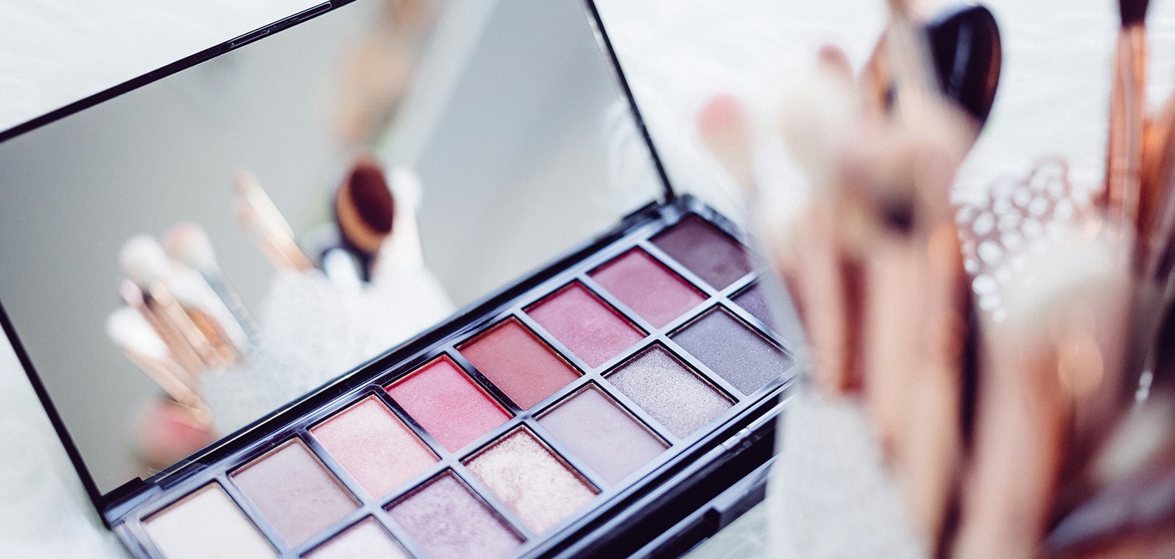 Eine Make-up-Palette mit verschiedenen Rosa- und Brauntönen, die sich im Spiegel reflektieren, umgeben von Make-up-Pinseln.