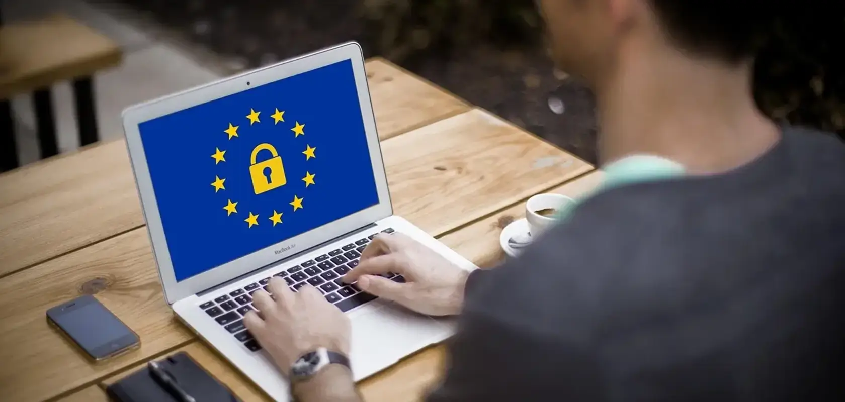 Person typing on laptop with EU flag and lock symbol on screen, sitting at a wooden table with a phone and cup nearby.