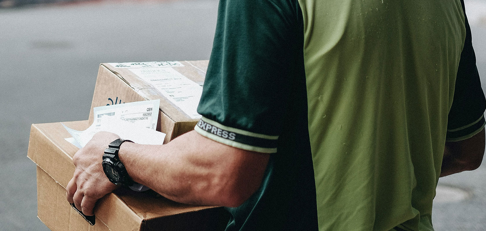 Lieferant in grüner Uniform, der gestapelte Kartons trägt und Versandpapiere in der Hand hält.