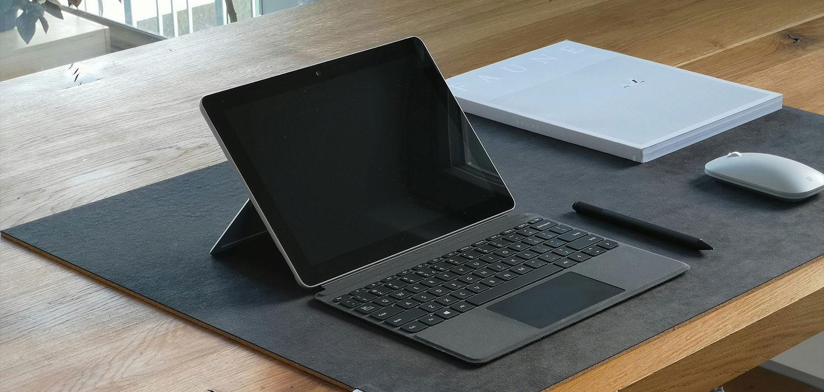 A tablet with a keyboard cover, stylus, and mouse on a wooden desk, accompanied by a closed white laptop.
