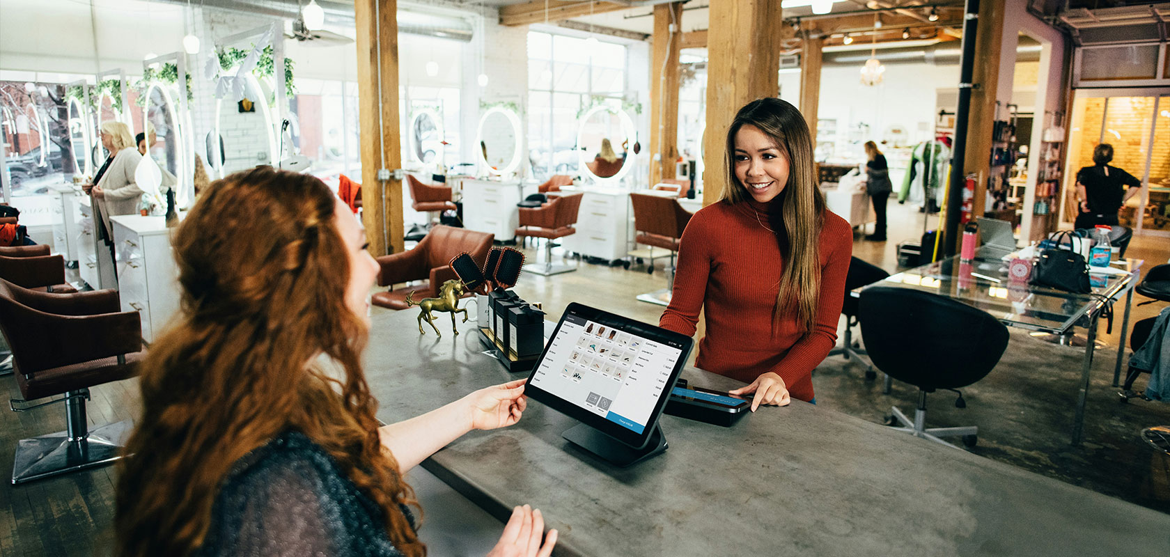 Two women chatting and smiling at the reception desk of a bustling, modern hair salon; customers can be seen in the background.