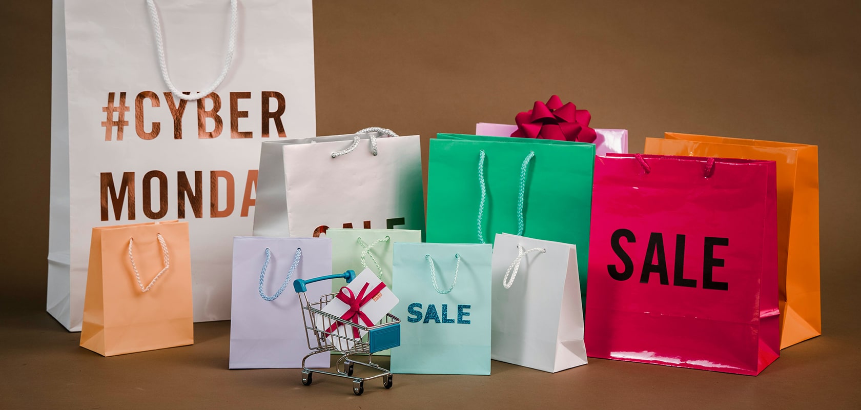 Bunte Einkaufstaschen mit den Aufschriften „SALE“ und „#CYBER MONDAY“ um einen Mini-Einkaufswagen mit einer kleinen Geschenkbox herum, auf braunem Hintergrund.