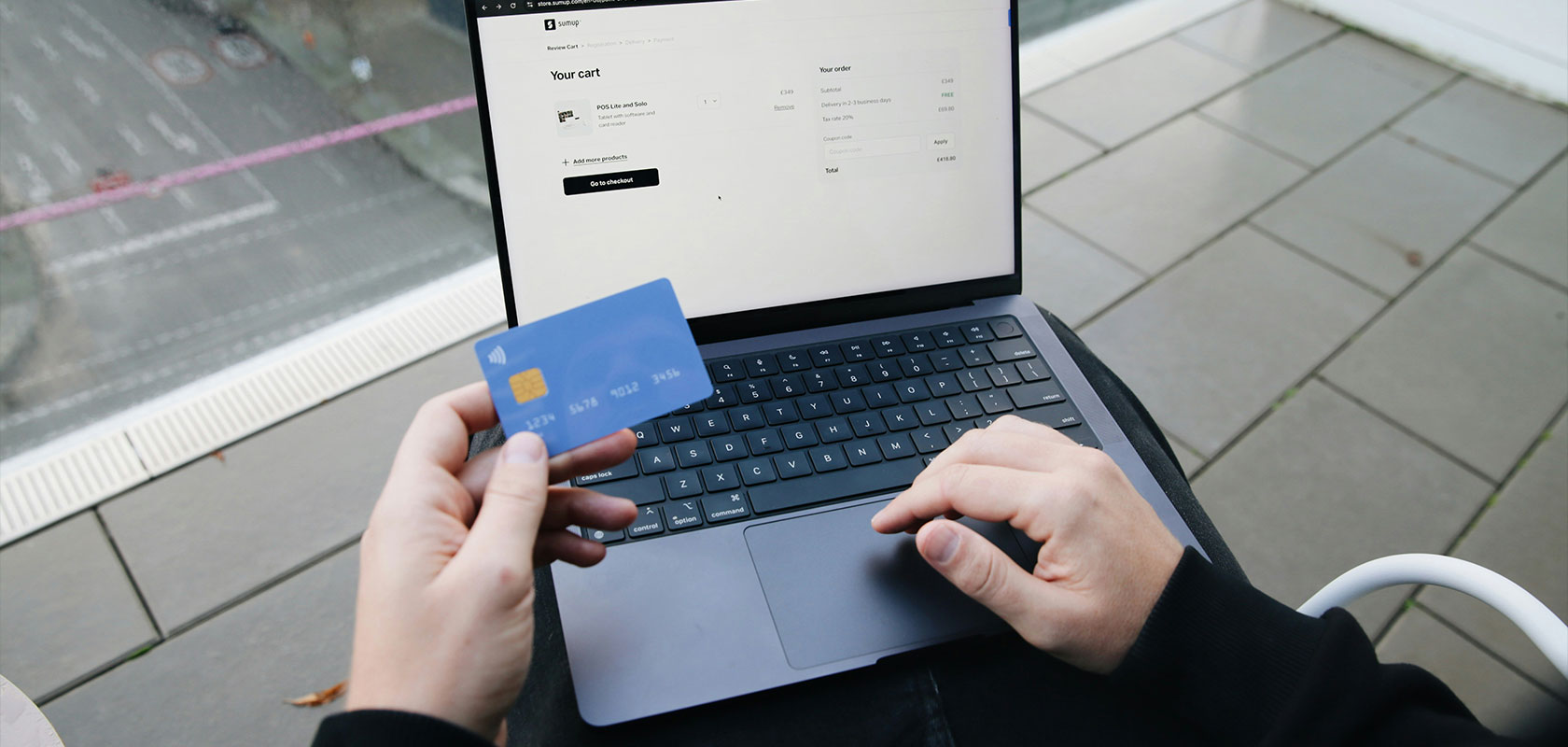 Person, die eine blaue Kreditkarte in der Hand hält, während sie einen Laptop benutzt, auf dessen Bildschirm ein Online-Warenkorb angezeigt wird.