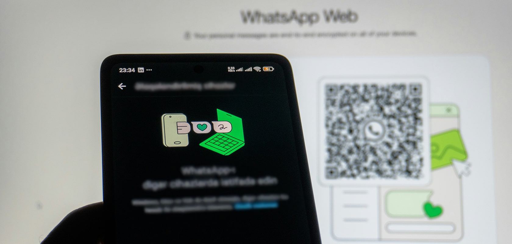 Smartphone mit WhatsApp Web-Anmeldebildschirm vor einem unscharfen Monitor, auf dem ein QR-Code angezeigt wird.