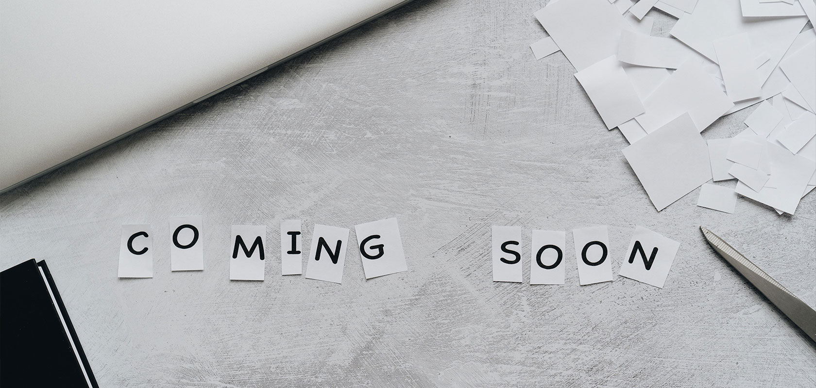 Der Schriftzug „COMING SOON“ aus ausgeschnittenen Papierbuchstaben auf einem hellgrauen Schreibtisch mit Office-Utensilien.