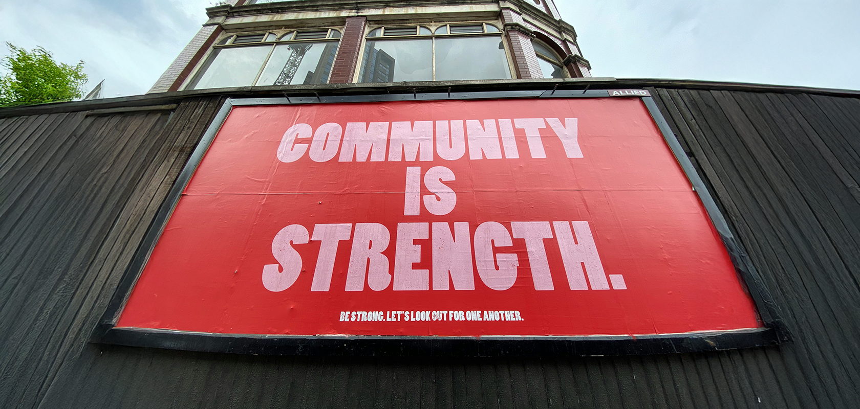 A red billboard with large white text reading "Community is Strength" on a black wooden wall, with a building facade above.