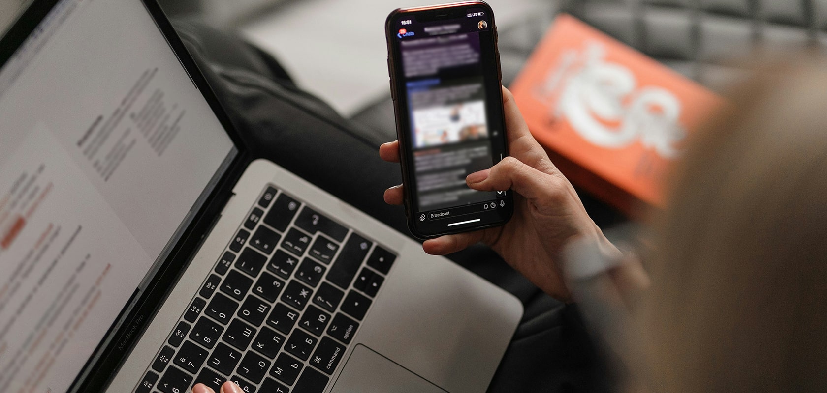 Person, die während der Arbeit am Laptop ein Smartphone benutzt, mit unscharfem Hintergrund, auf dem eine Kiste und ein Sofa zu sehen sind.