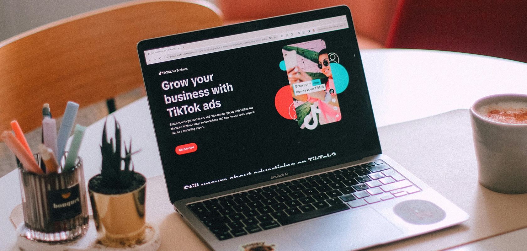Auf einem Tisch liegt ein geöffneter Laptop mit einer Webseite mit dem Titel „Wachsen Sie mit TikTok-Anzeigen“, daneben stehen Stifte in einem Stifthalter, eine kleine Pflanze und eine Kaffeetasse.
