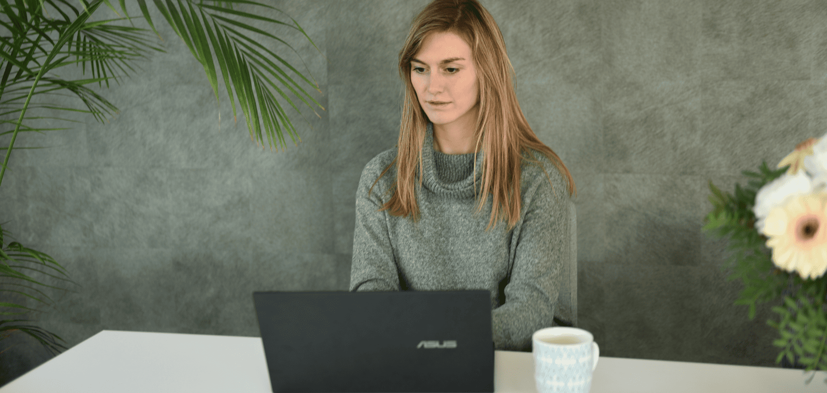 Woman in a green sweater works on a laptop at a white desk, with a mug and flowers nearby, set against a gray background with plants.