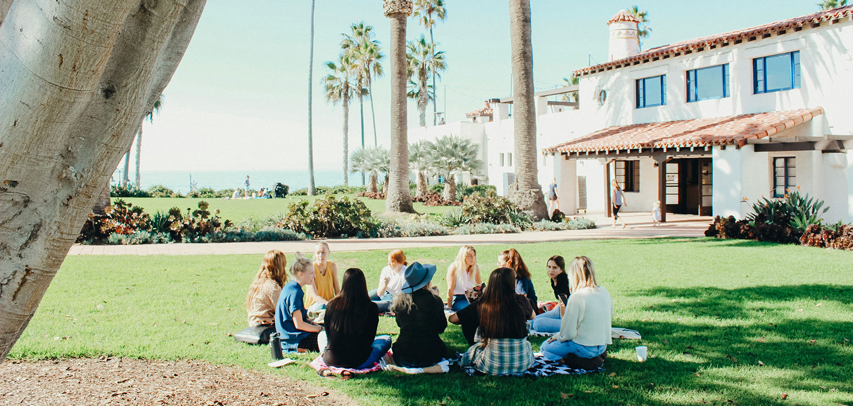 Eine Gruppe von Menschen sitzt im Kreis auf einer Rasenfläche in der Nähe eines großen weißen Gebäudes, im Hintergrund sind Palmen und das Meer zu sehen.