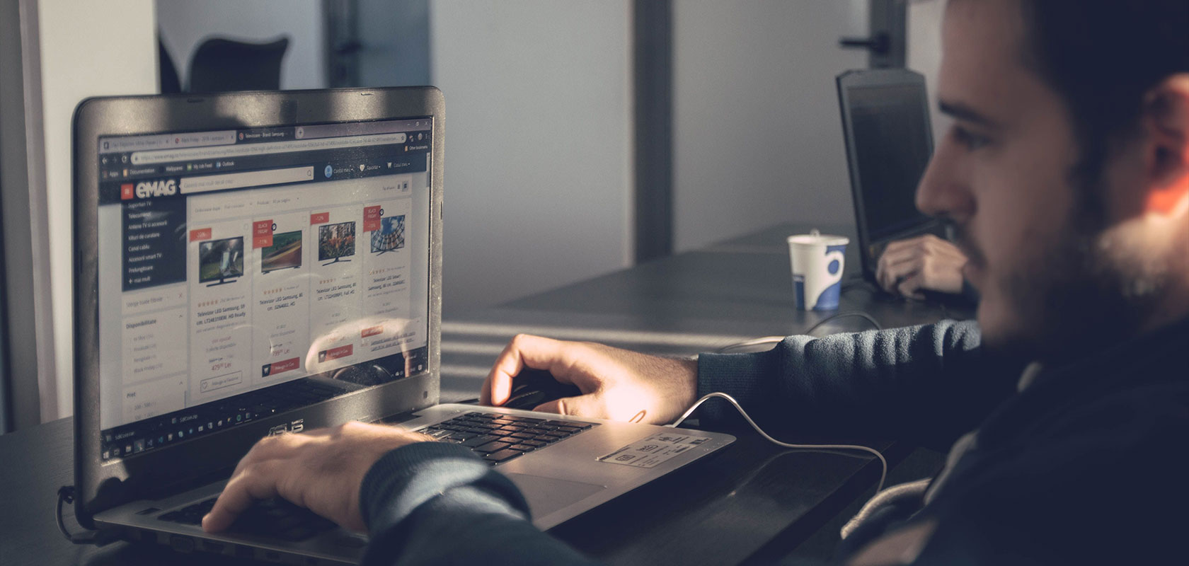 Eine Person sitzt an einem Schreibtisch und surft mit einem Laptop auf einer Website, auf der Artikel zum Thema Auto zu sehen sind; im Hintergrund steht eine Kaffeetasse.