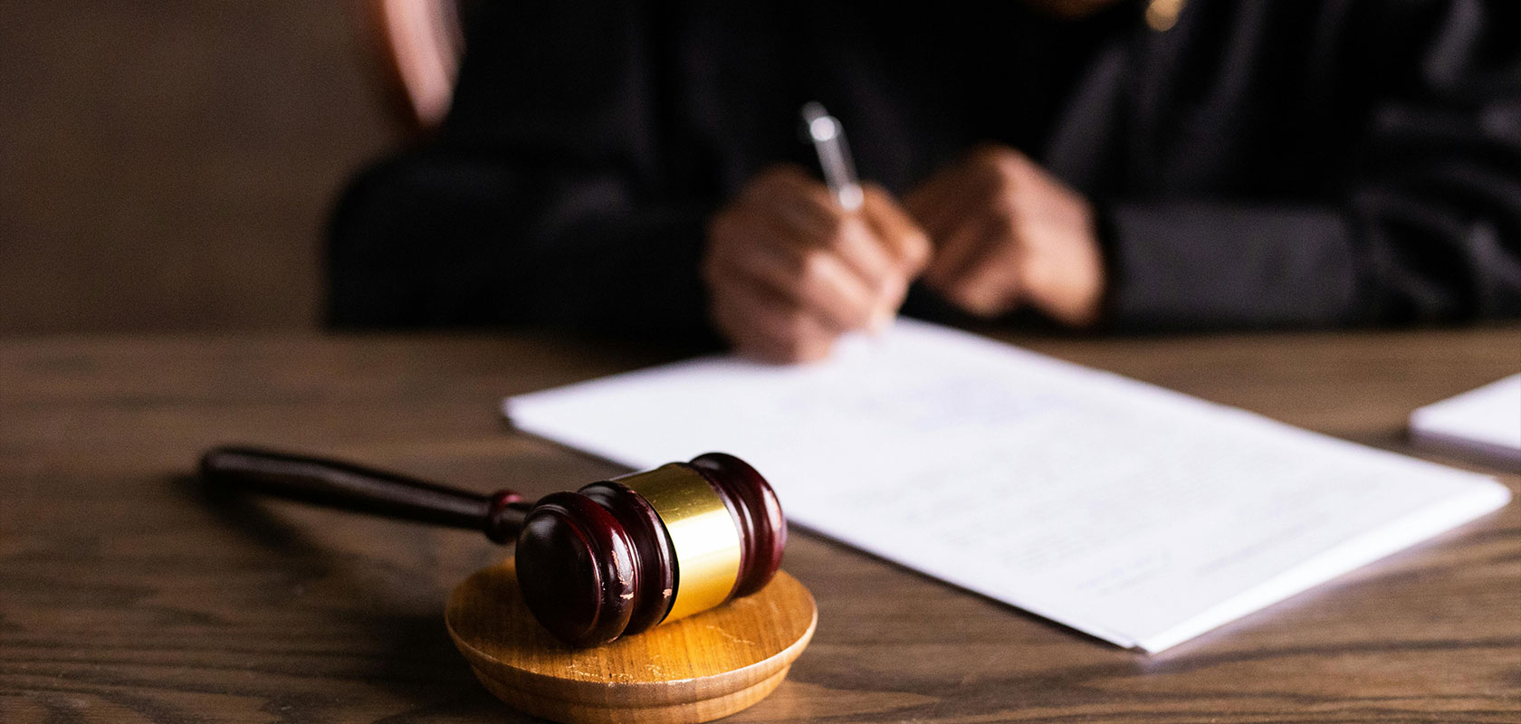 Ein Richter schreibt an einem Schreibtisch auf ein Blatt Papier, im Vordergrund ein Richterhammer, der Gerichtsverfahren und Entscheidungsfindung symbolisiert.