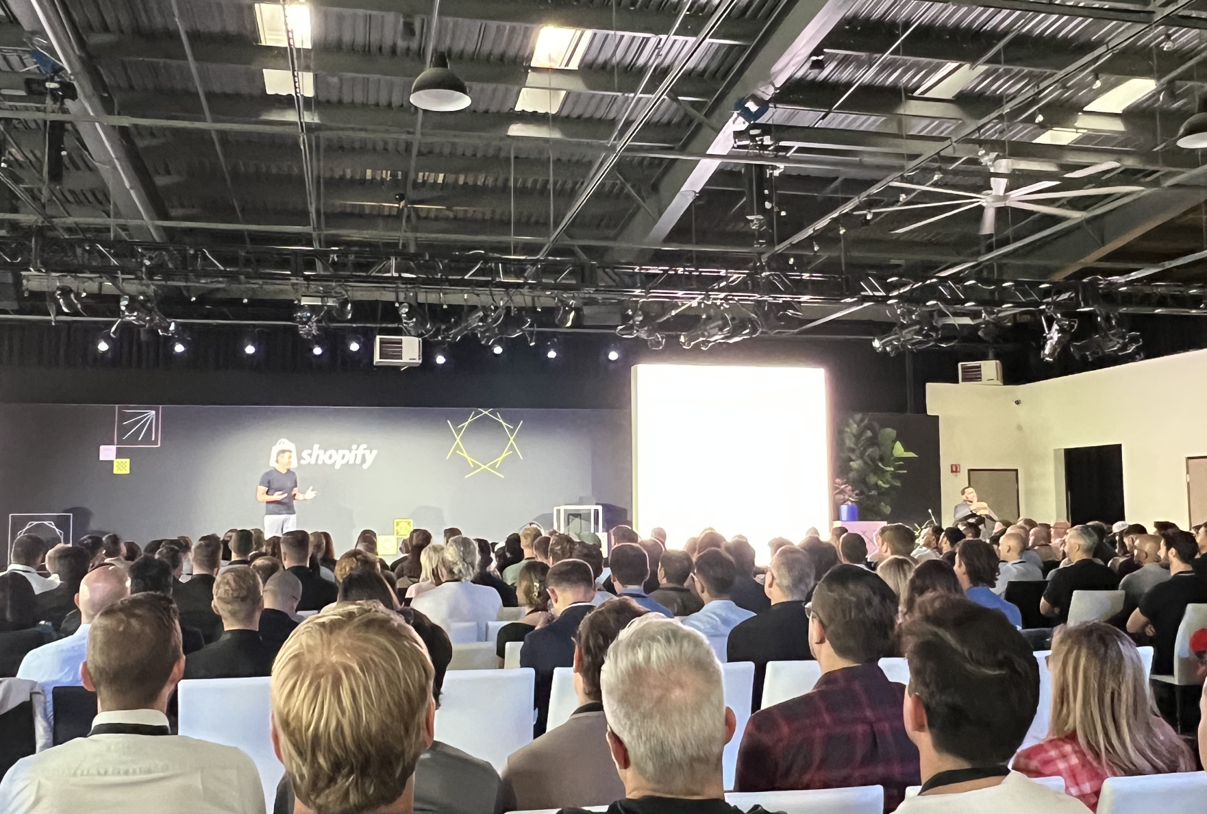 Ein Redner hält vor einem großen Publikum bei einer Shopify-Veranstaltung in einem modernen Konferenzsaal mit industrieller Decke und heller Beleuchtung eine Präsentation.