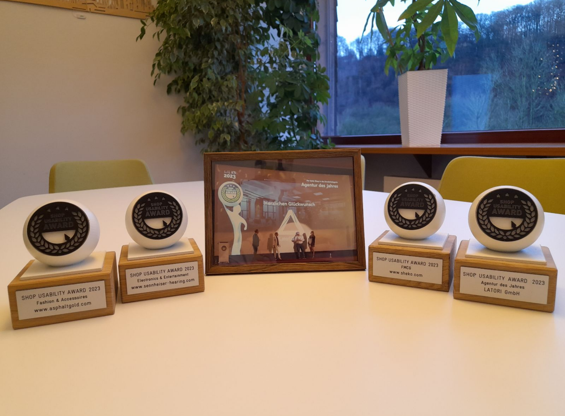 Fünf Auszeichnungen auf einem Tisch am Fenster, vier mit runden Plaketten und eine gerahmt, draußen sind Bäume zu sehen.