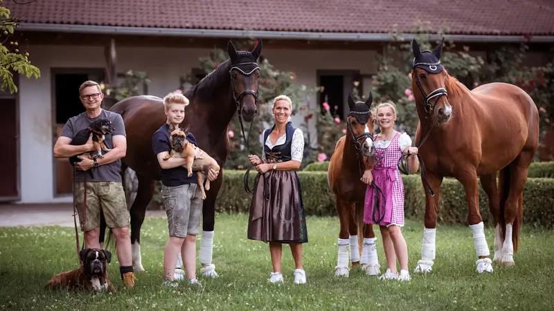 Eine Familie mit zwei Hunden und zwei Pferden steht lächelnd auf einer Wiese vor einem Stall. Die Pferde tragen Zaumzeug und Beinschlaufen.