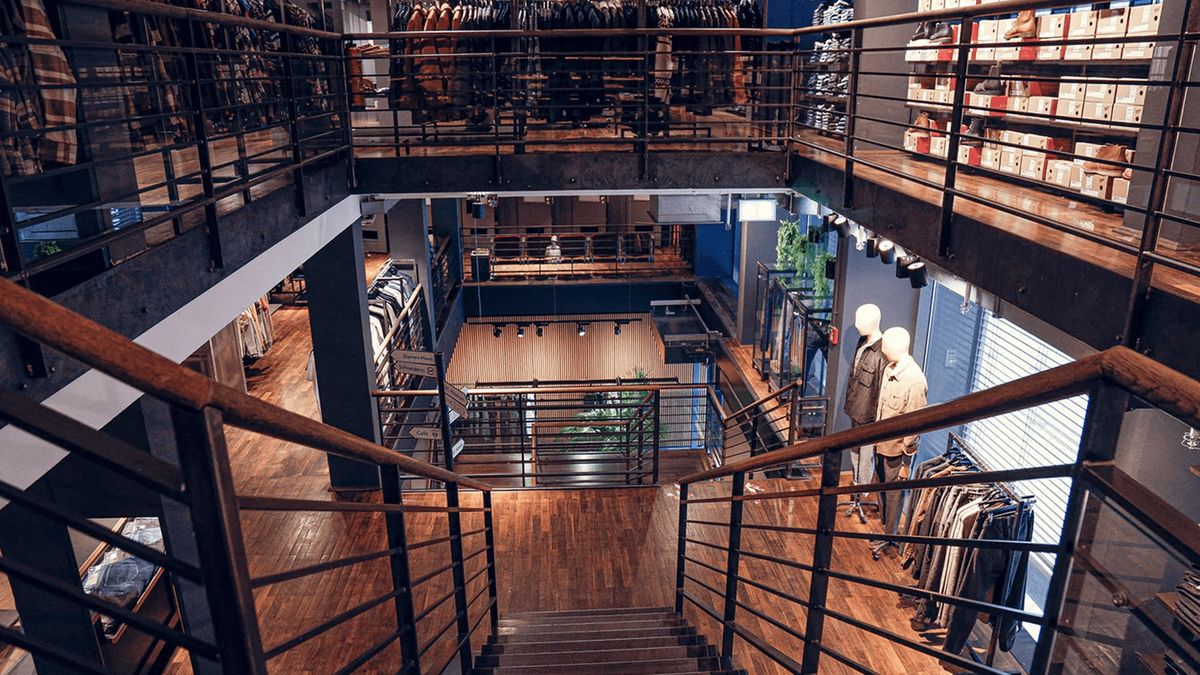 Innenraum eines modernen Bekleidungsgeschäfts mit Holzböden, Metallgeländern, Schaufensterpuppen und ordentlich angeordneter Kleidung in Regalen und Gestellen.