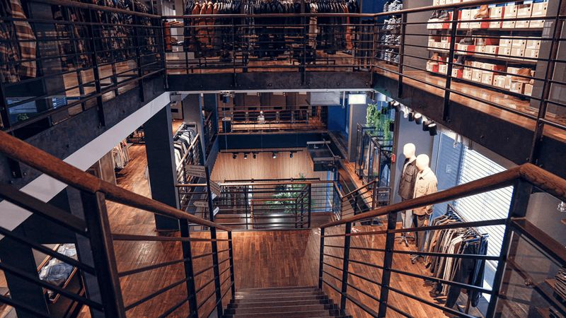 Innenraum eines modernen Bekleidungsgeschäfts mit Holzböden, Metallgeländern, Schaufensterpuppen und ordentlich angeordneter Kleidung in Regalen und Gestellen.