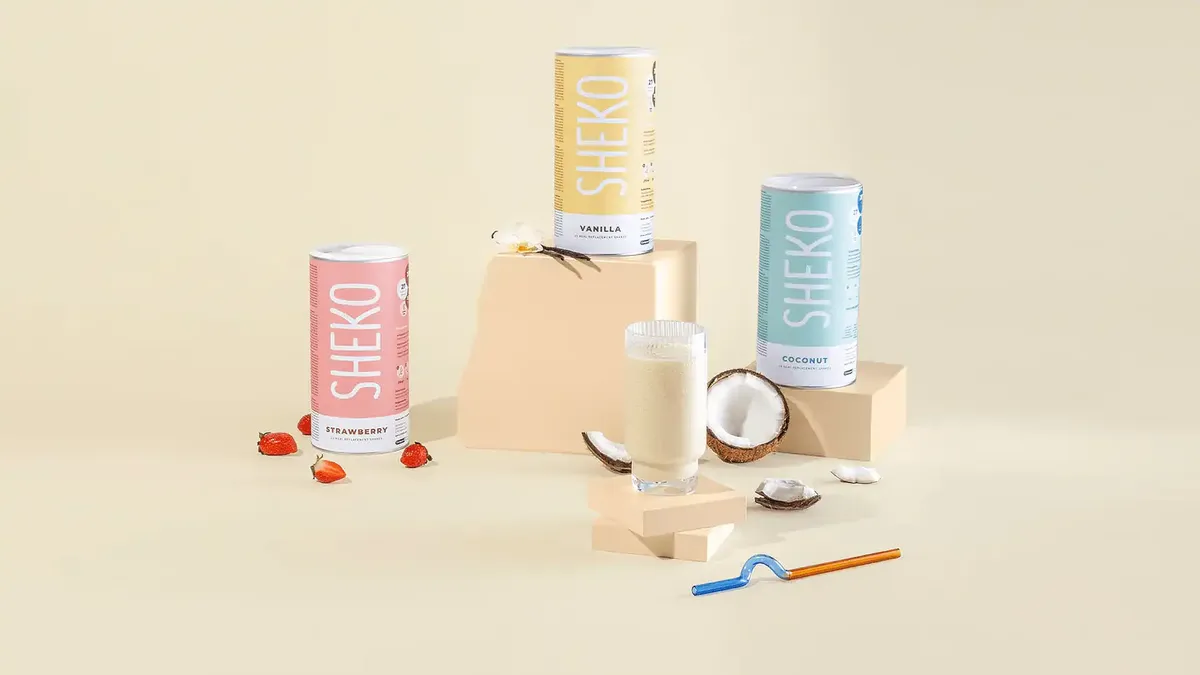 Drei Dosen SHEKO-Proteinshake in den Geschmacksrichtungen Erdbeere, Vanille und Kokosnuss, mit einem Glas Shake und Kokosnuss-Stücken auf einem beigen Hintergrund.