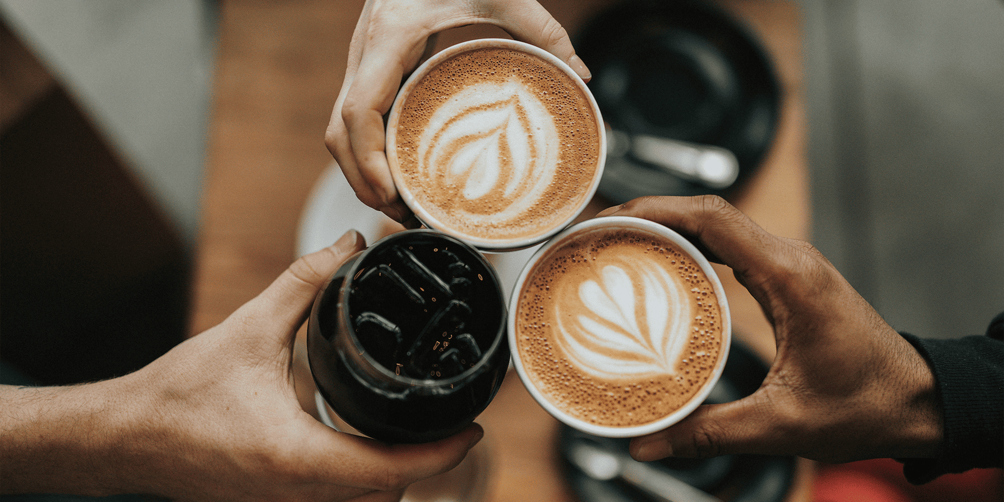 Drei Hände halten Getränke über einem Tisch: zwei Latte Macchiatos mit Latte-Art und ein Glas Eiskaffee. Darunter sind schwarze Untertassen zu sehen.