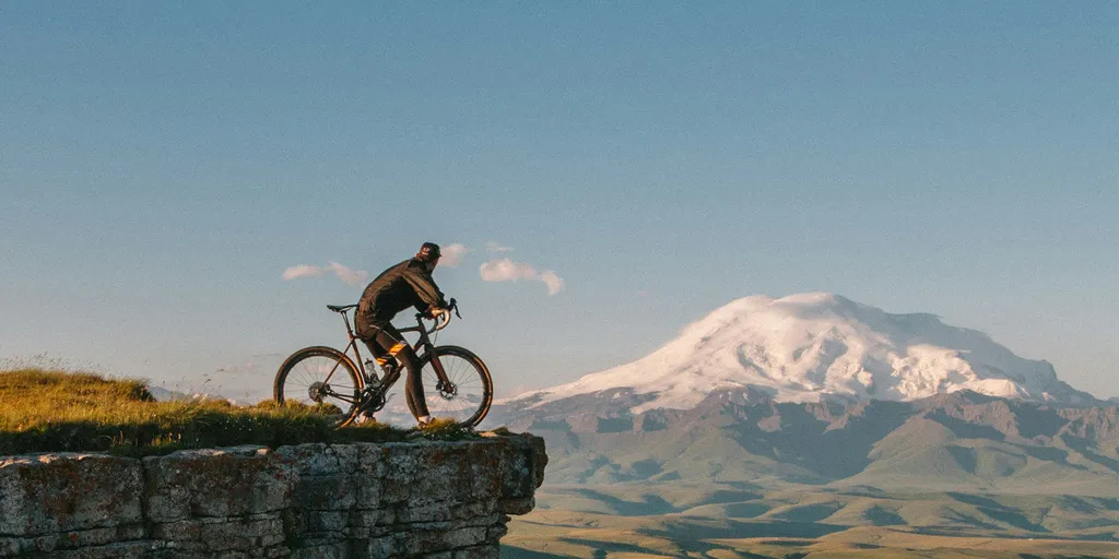 Ein Radfahrer steht am Rand einer felsigen Klippe und blickt auf eine weite Landschaft mit einem schneebedeckten Berg unter einem strahlend blauen Himmel.