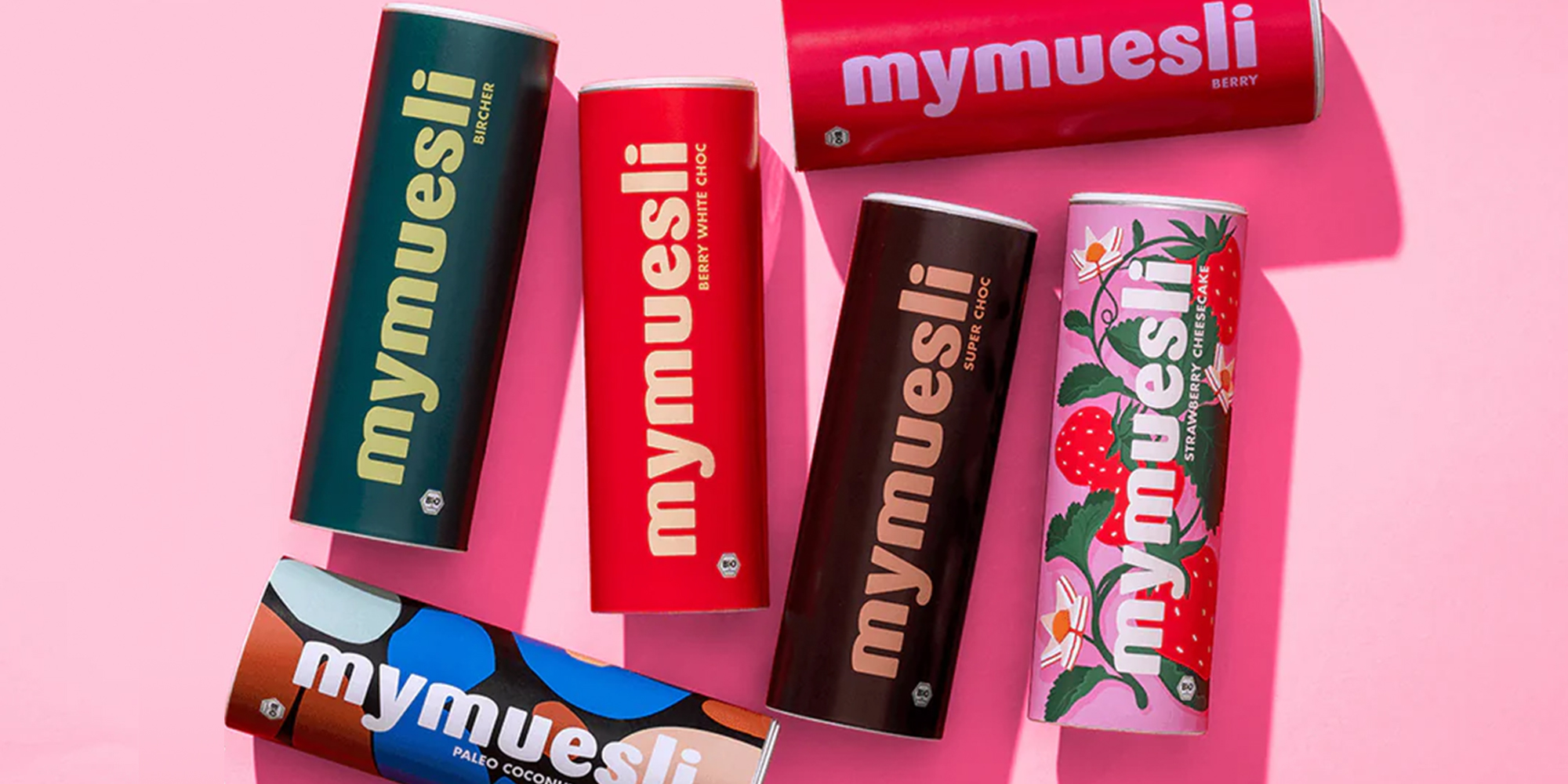 Bunte zylindrische Mymuesli-Behälter auf rosa Hintergrund mit verschiedenen Designs und Geschmacksrichtungen.