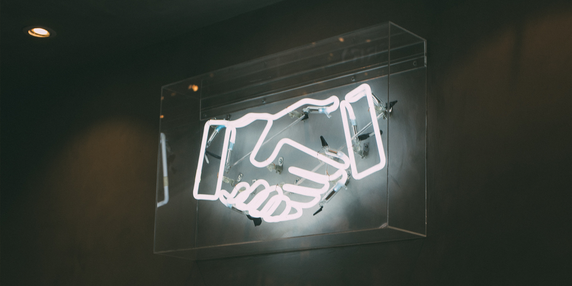 Neon sign of two hands shaking, mounted in a transparent case against a dark wall, with a single ceiling light.