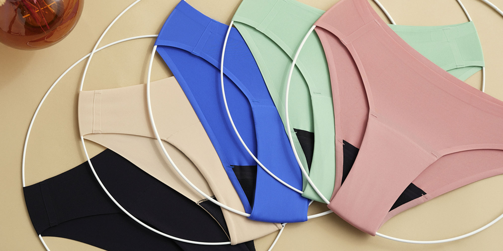 Fünf Paar farbenfrohe Damenunterwäsche, angeordnet innerhalb sich überlappender weißer Kreise auf einer beigen Fläche, neben einer Vase mit Trockenblumen.