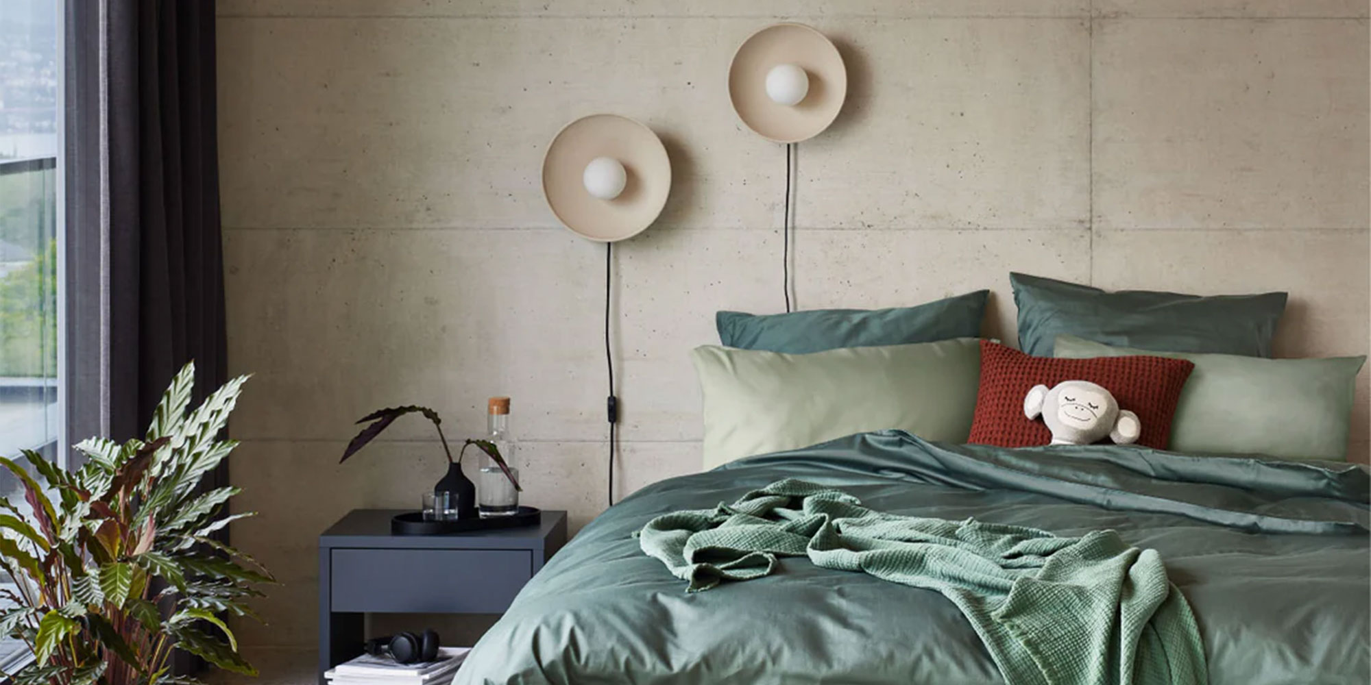 Gemütliches Schlafzimmer mit grüner Tagesdecke, dekorativen Kissen, Wandlampen, einem Nachttisch mit Pflanzen und einem mit Vorhängen verdeckten Fenster.