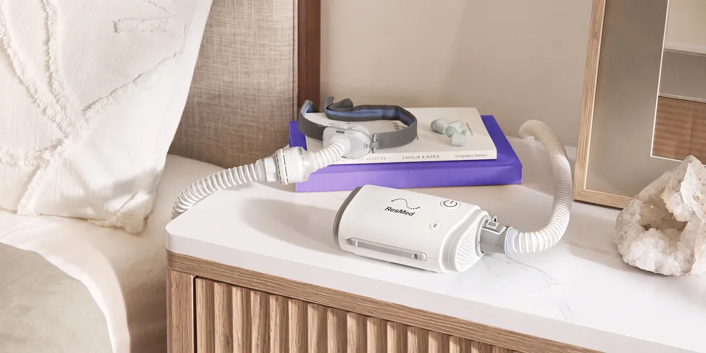 Ein CPAP-Gerät auf einem Nachttisch mit einer Gesichtsmaske, einem Schlauch und einem violetten Buch neben einem beigen Bett. Außerdem ist eine Kristallverzierung zu sehen.