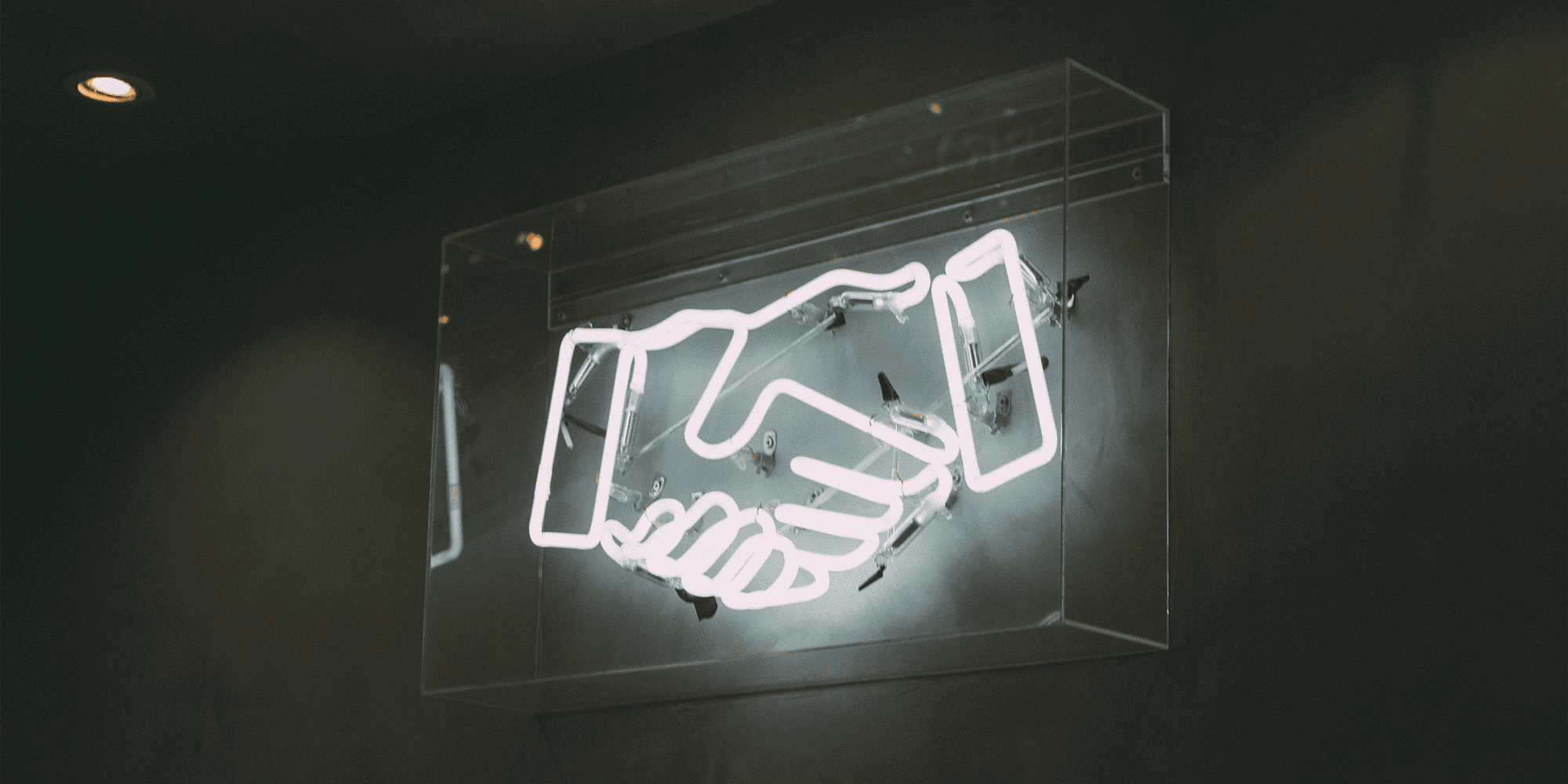 A glowing neon sign in the shape of two hands clasped together, enclosed in a transparent acrylic case on a dark wall.