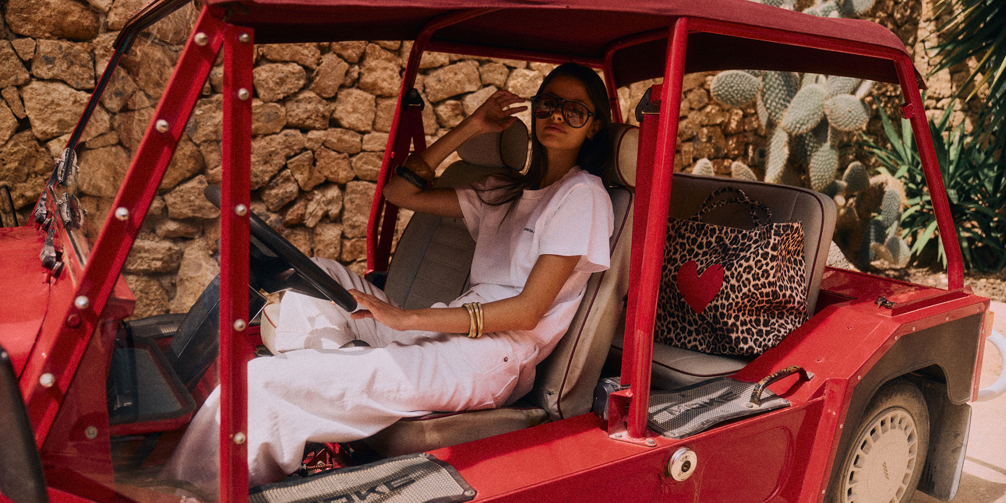 Eine Frau mit Sonnenbrille sitzt in einem roten Cabrio, das neben einer Steinmauer und Kakteen geparkt ist, mit einer Tasche mit Leopardenmuster auf dem Sitz.