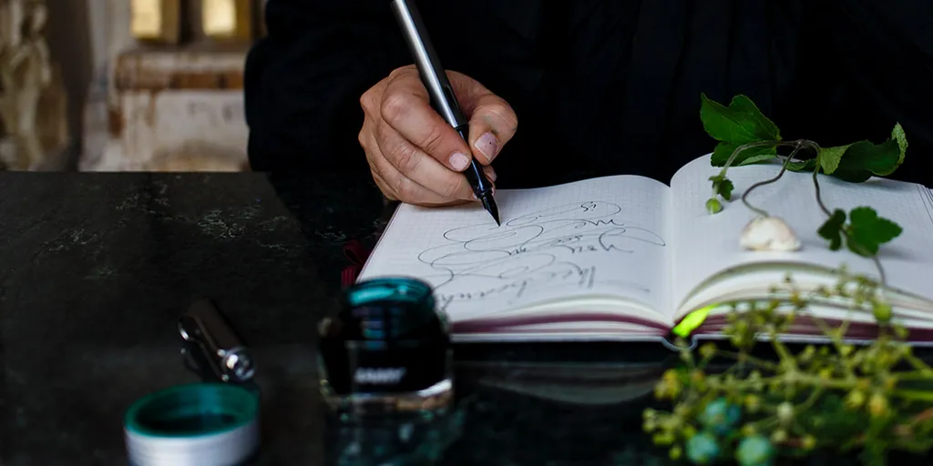 Eine Person, die mit einem Stift in ein Notizbuch schreibt, umgeben von Grünzeug und einem Tintenfass auf einer dunklen Fläche.