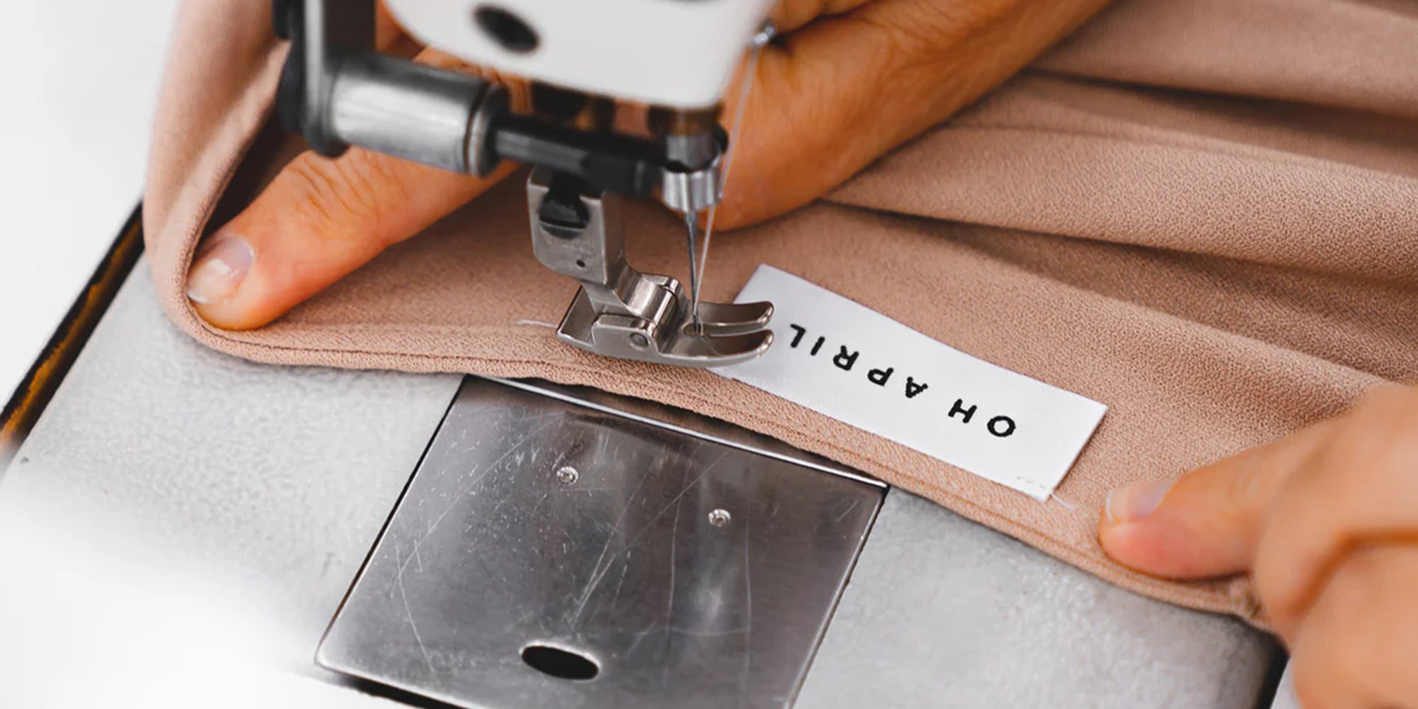 Hände, die einen beigen Stoff mit einer Nähmaschine nähen und ein Etikett mit der Aufschrift „OH APRIL“ anbringen.