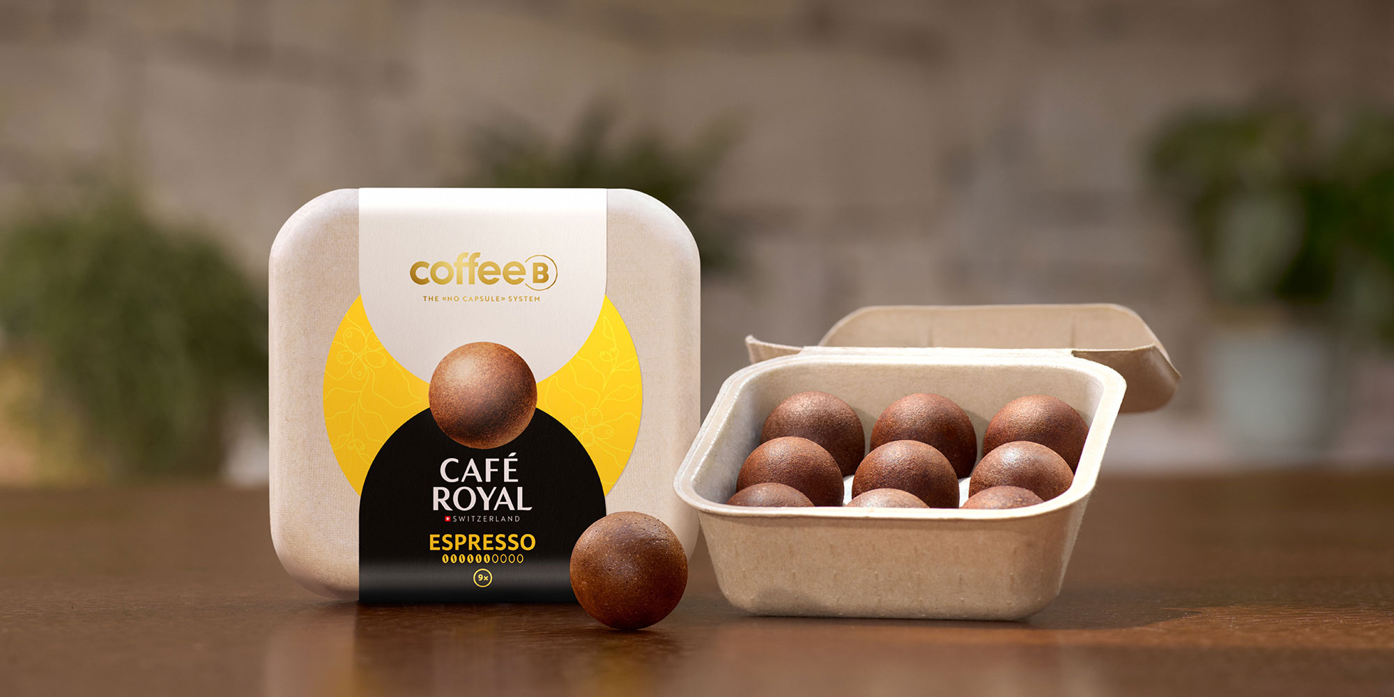 CoffeeB Café Royal Espresso pods in a square container, with one pod outside, on a wooden surface. Background is softly blurred.