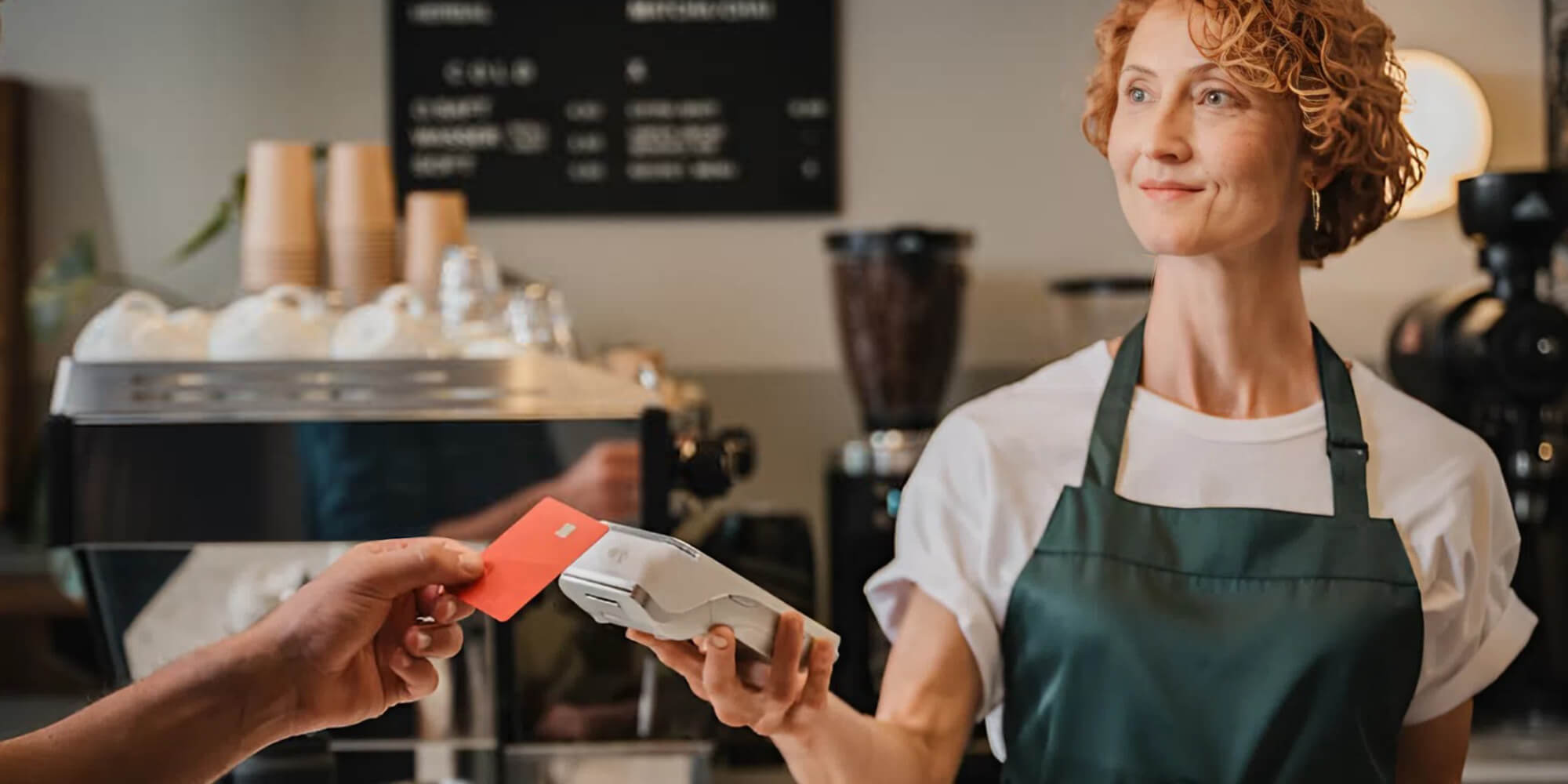 Ein Barista in einer grünen Schürze hält einen Kartenleser in der Hand, während ein Kunde eine rote Karte vorzeigt. Im Hintergrund ist eine Kaffeetheke zu sehen.