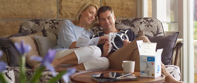 Ein Paar sitzt auf einem gemütlichen Sofa und baut ein Gerät zusammen, während es die Anleitung liest; im Vordergrund stehen ein Couchtisch und Blumen.