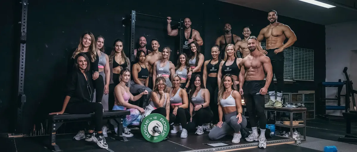 Eine Gruppe von fitten Personen posiert in einem Fitnessstudio in Trainingskleidung. Einige sitzen, andere stehen, mit Fitnessgeräten im Hintergrund.