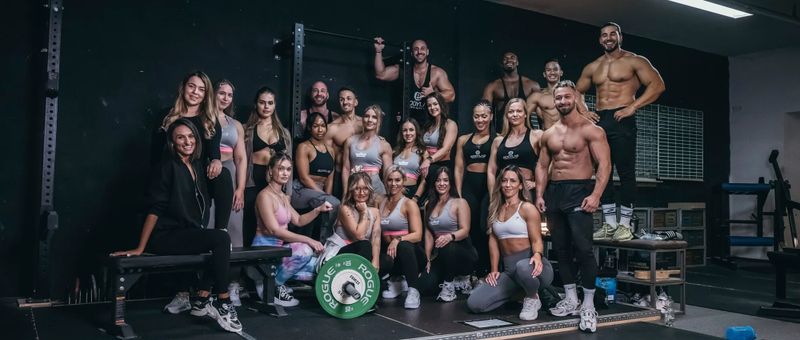 Eine Gruppe von fitten Personen posiert in einem Fitnessstudio in Trainingskleidung. Einige sitzen, andere stehen, mit Fitnessgeräten im Hintergrund.