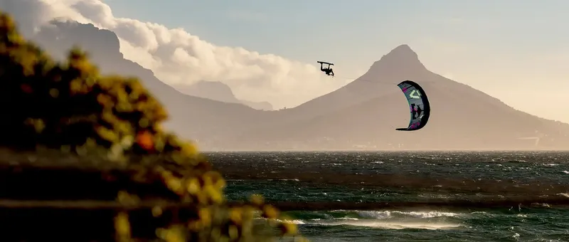 Eine Person, die bei Sonnenuntergang unter einem teilweise bewölkten Himmel vor einer dramatischen Bergkulisse über ein windiges Meer kitesurft.