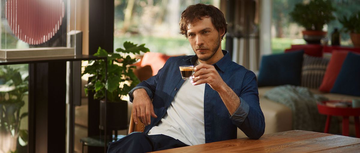 A man in a blue jacket sits at a wooden table, holding an espresso, with plants and a cozy interior in the background.