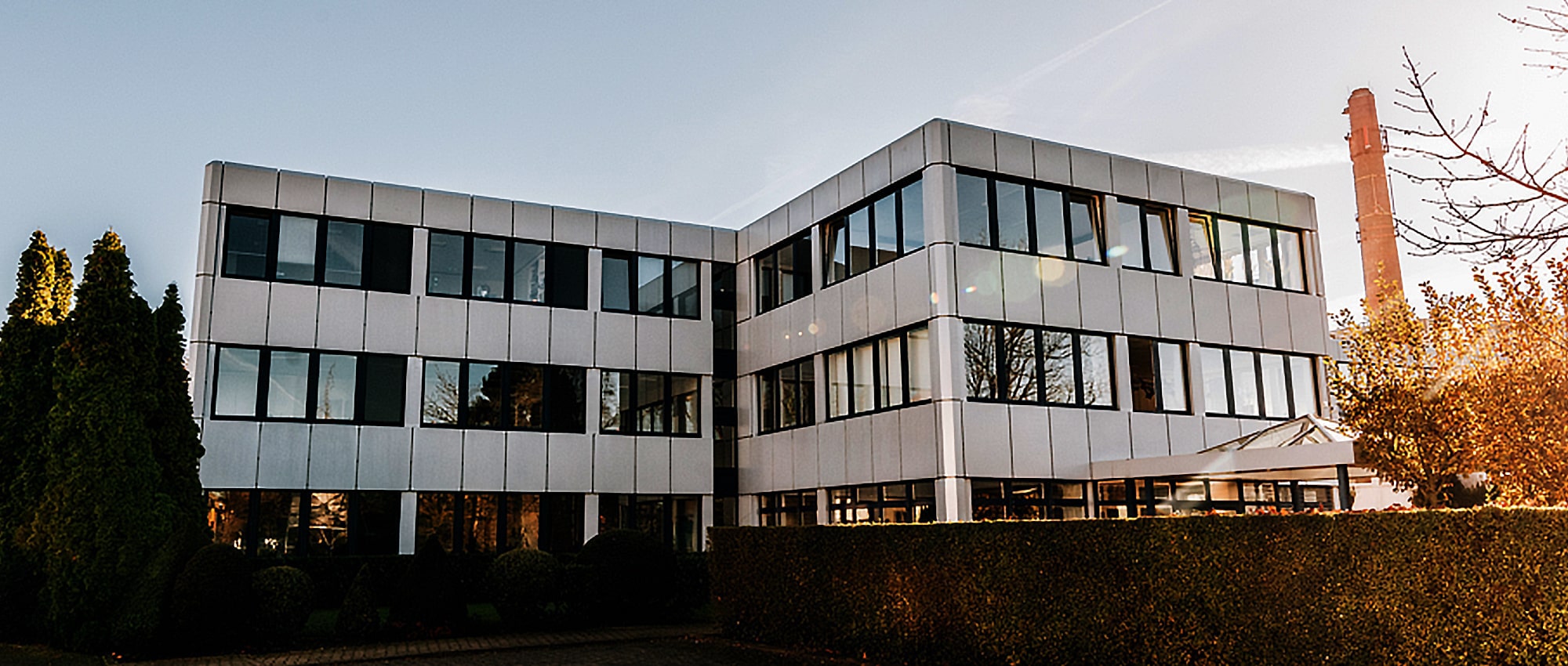 Modernes dreistöckiges Bürogebäude mit großen Fenstern, umgeben von Bäumen und einer Hecke, unter einem strahlend blauen Himmel.
