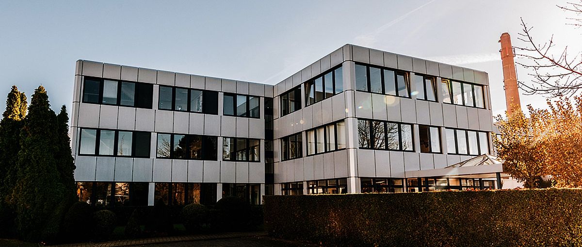 Modernes dreistöckiges Bürogebäude mit großen Fenstern, umgeben von Bäumen und einer Hecke, unter einem strahlend blauen Himmel.