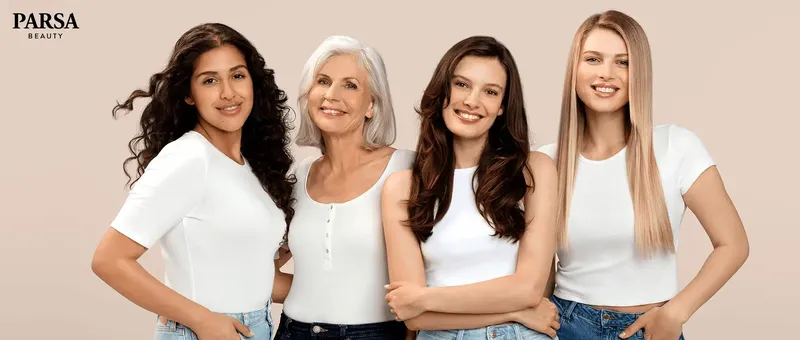 Vier Frauen unterschiedlichen Alters lächeln, tragen weiße Oberteile und Jeans und stehen vor einem beigen Hintergrund.