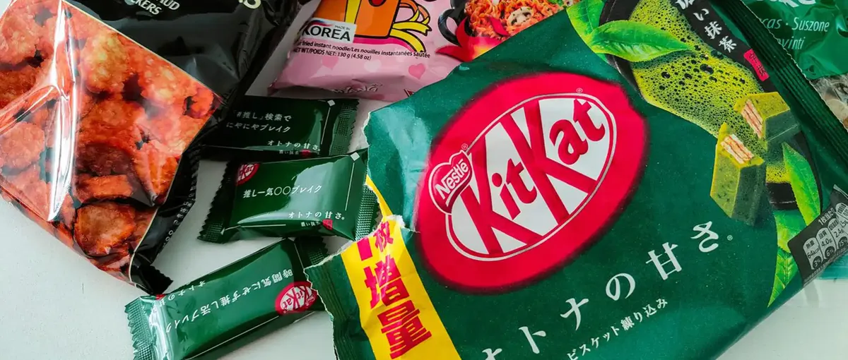 Verschiedene asiatische Snacks, darunter ein Kit Kat mit grünem Tee, verschiedene Süßigkeiten und Chips, ausgestellt auf einer weißen Fläche.