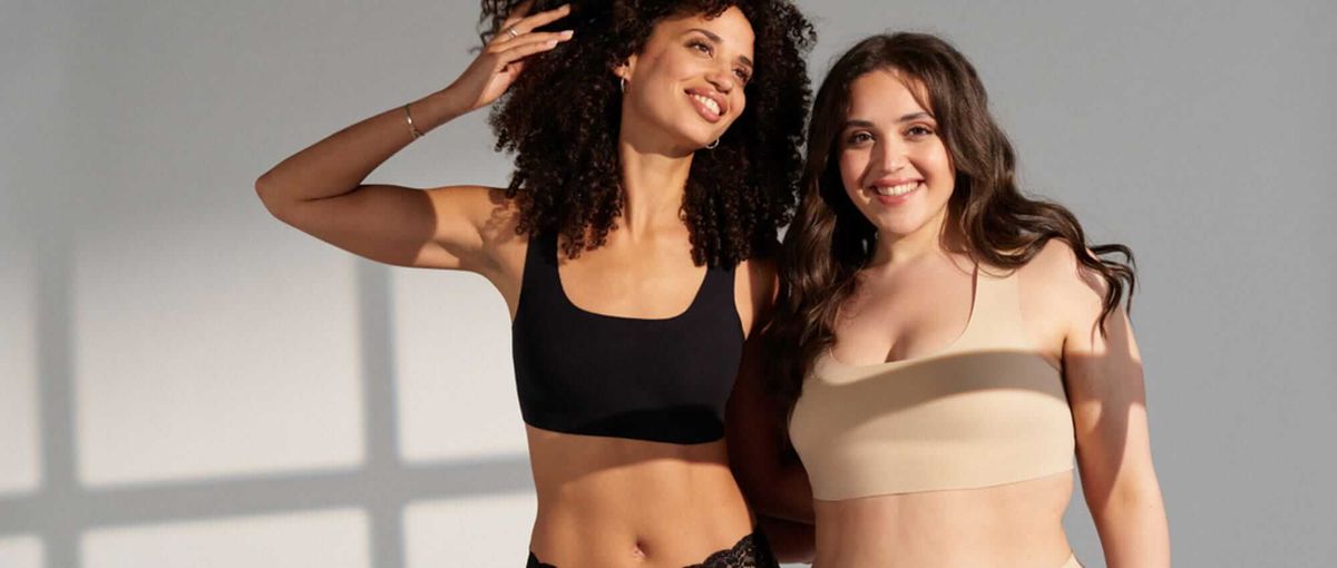 Zwei Frauen lächeln in einem hell erleuchteten Raum und tragen schwarze und beige Unterwäsche. Sonnenlicht wirft Schatten auf die Wand hinter ihnen.