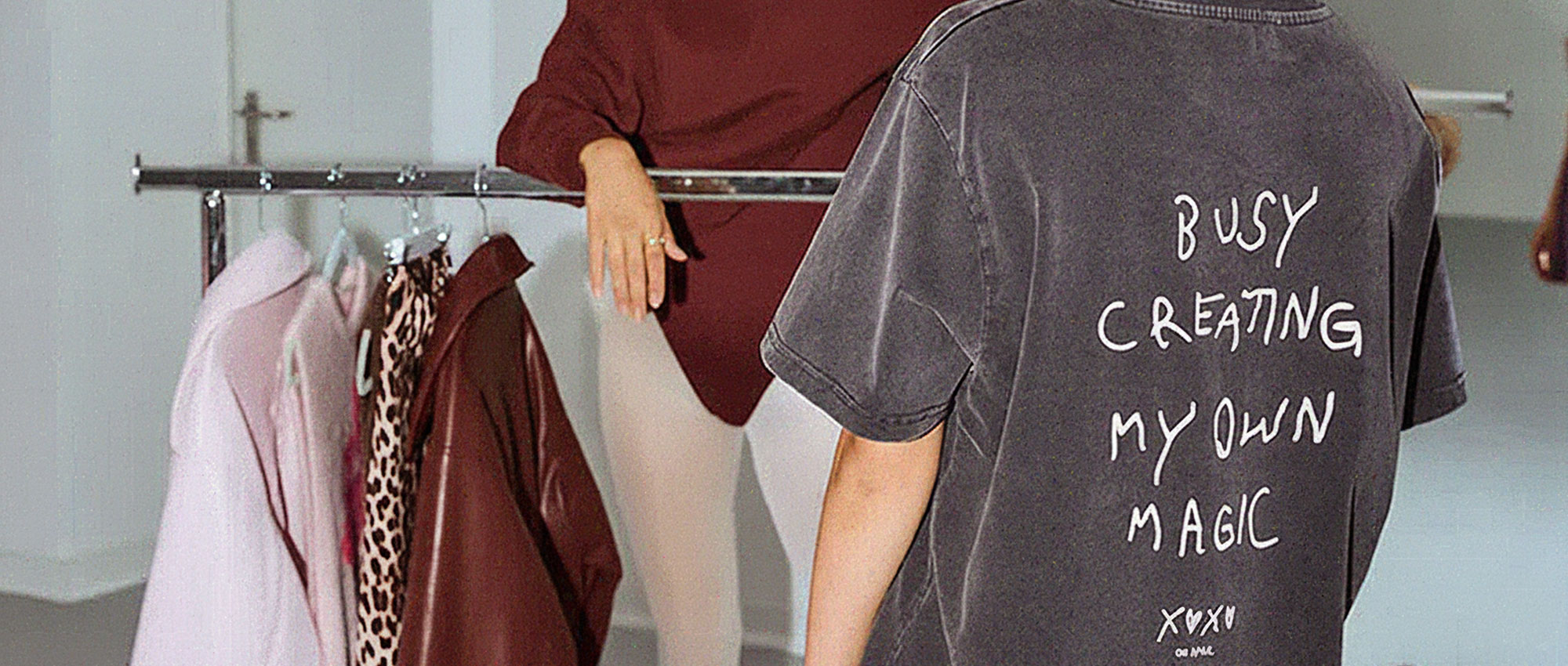 Person mit einem verblassten schwarzen T-Shirt mit der Aufschrift „Busy creating my own magic” (Beschäftigt damit, meine eigene Magie zu erschaffen) auf dem Rücken, stehend neben einem Kleiderständer mit Mänteln und einer Person in Burgunderrot.