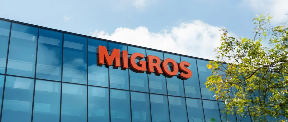 Das Bild zeigt die Außenansicht eines Gebäudes mit einem großen roten ‚MIGROS‘-Schriftzug vor Glasfenstern, im Hintergrund ein blauer Himmel und Baumblätter.